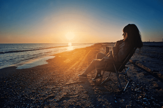 Woman sitting in beach chair wearing coat and holding coffee cup. Looking out at waves.