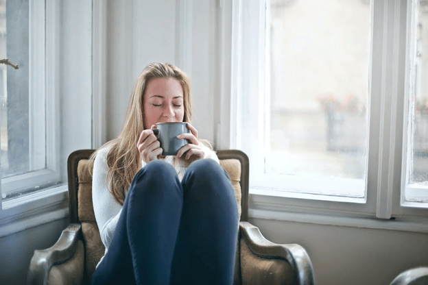A young woman with long blond hair wearing blue slacks and a white sweater sits in a tan and wood trimmed antique chair in a corner of a room with windows on both sides. Her eyes are closed and she holds a greyish colored coffee mug which she appears to be savoring..