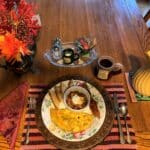 A hearty Fall breakfast of stuffed omelet baked apples and bacon sits on a festive plate and metal charger. Table setting has red and orange flowers in a silver pot, a red coffee mug and cream colored carafe with a glass condiment tray above the plate.