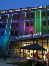Modern building with blue, red and green stripes of spotlight on it. Top of building sign reads Fox Cities Building for the Arts, while sign over small attached glass building reads The Trout Museum of Art.
